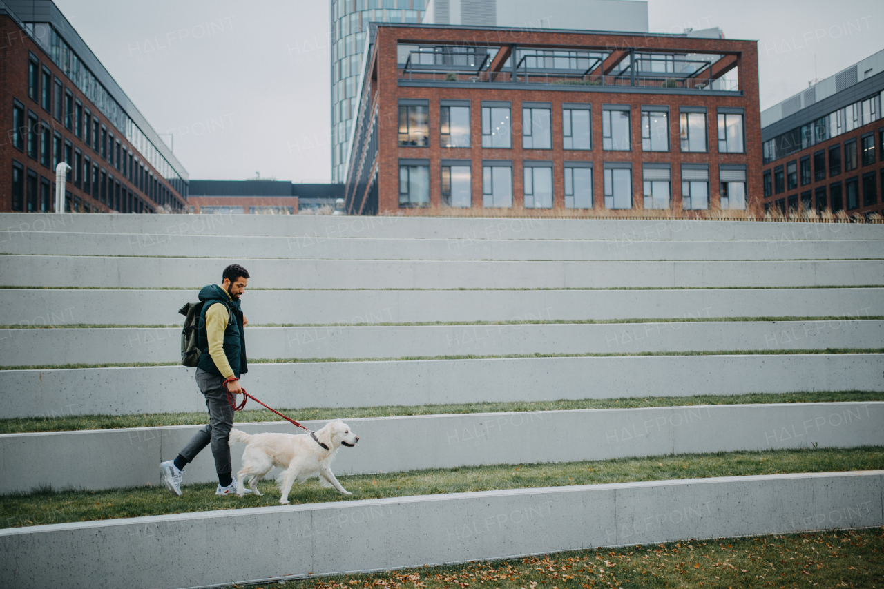 A happy young man walking his dog outdoors in city during grey autumn day.