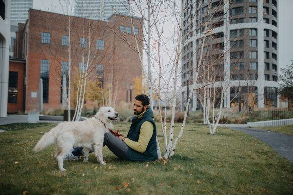 Happy young man stroking his dog outdoors in city park, during a cold autumn day.