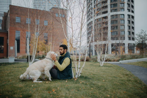 A happy young man sitting on grass with his dog outdoors in town.