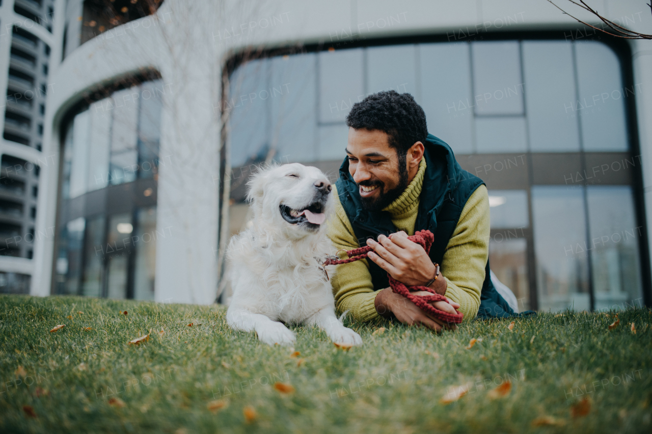 A happy young man lying on grass with his dog outdoors in town.