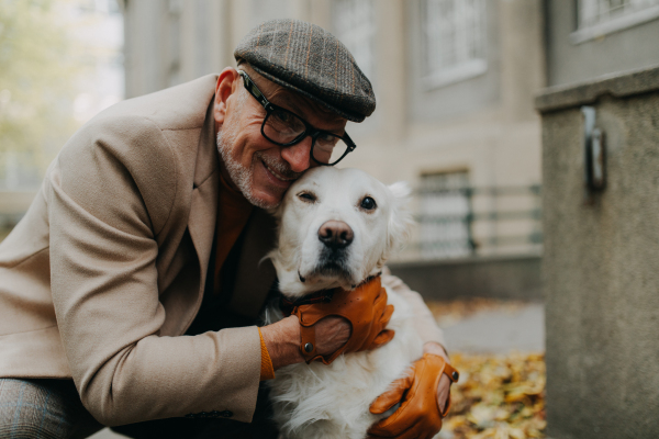 A happy senior man looking at camera and embracing his dog during walk outdoors in city.