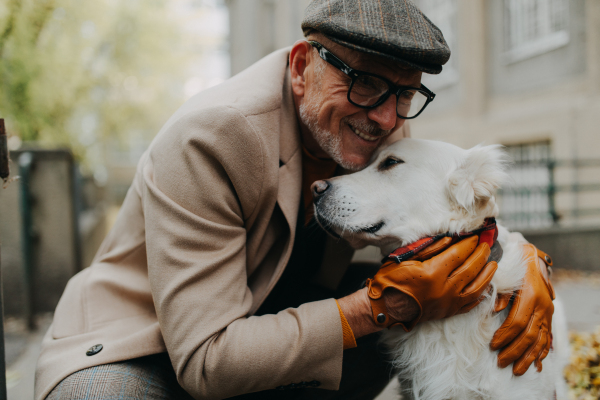 A happy senior man sitting on bench and embracing his dog outdoors in park in city.