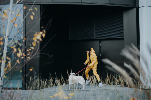 A happy young man walking his dog outdoors in city during autumn day.