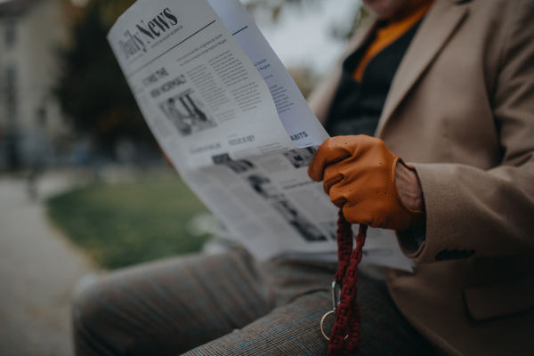 A senior man sitting on bench and reading newspaper outdoors in city park.