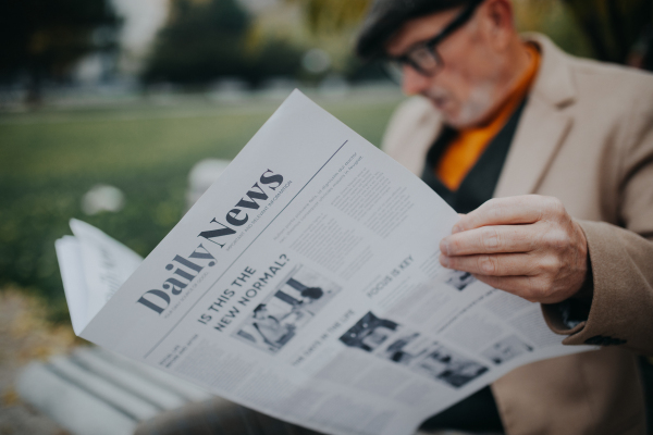 A happy senior man sitting on bench and reading newspaper outdoors in city park.