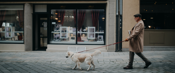 A side view of elegant senior man with take away coffee walking his dog outdoors in city in winter.