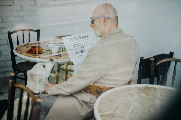 Happy senior man sitting in a cafeteria with his dog and reading newspaper.