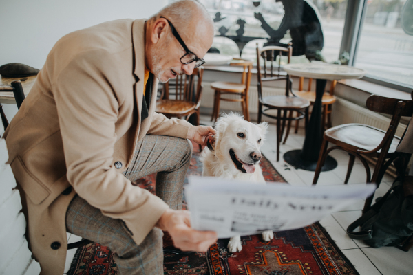 A happy senior man sitting in cafeteria and reading newspaper with his dog.