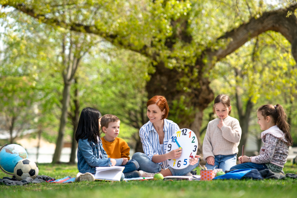 A teacher with small children sitting outdoors in city park, learning group education concept.