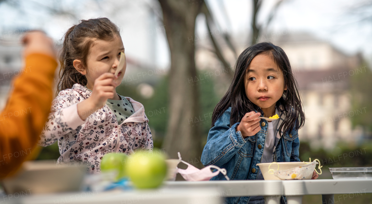 Happy small children eating lunch outdoors in city park, learning group education and coronavirus concept.