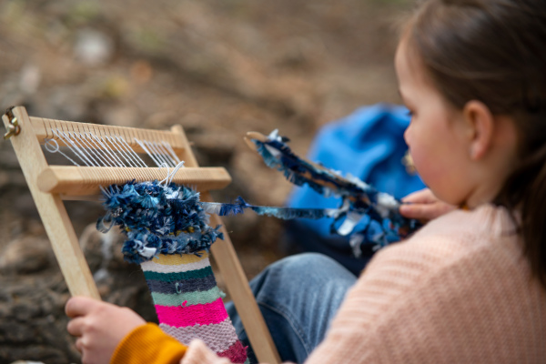 Happy small girl with hand loom sitting outdoors in city park, learning group education concept.