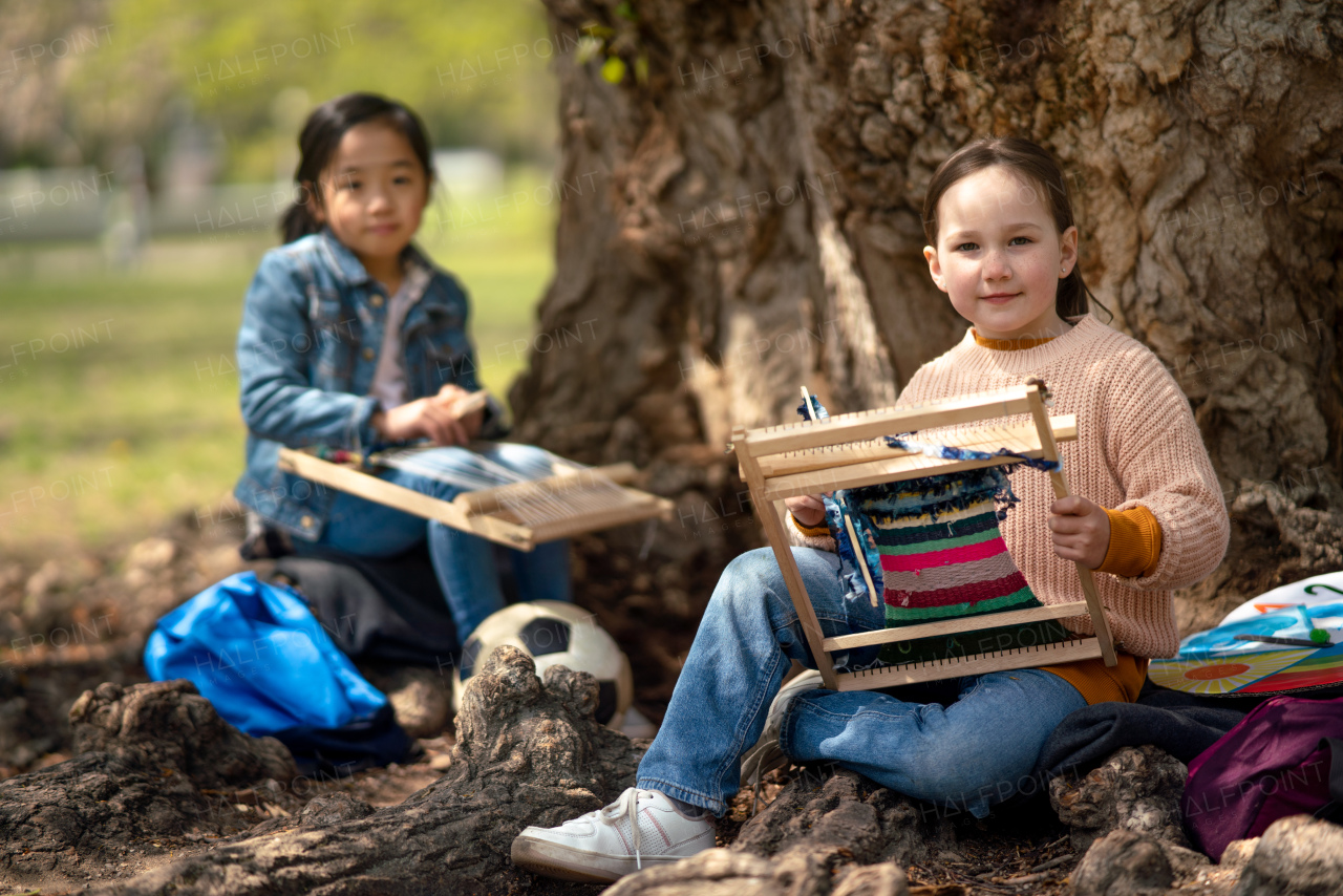 Portrait of small children with hand loom sitting outdoors in city park, learning group education concept.