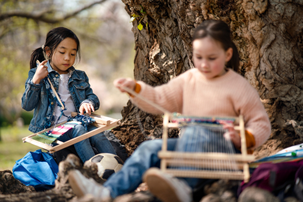 Portrait of small children with hand loom sitting outdoors in city park, learning group education concept.