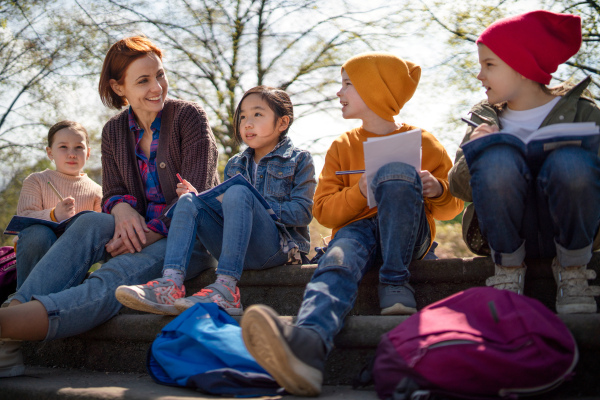 A teacher with small children sitting outdoors in city park, learning group education concept.