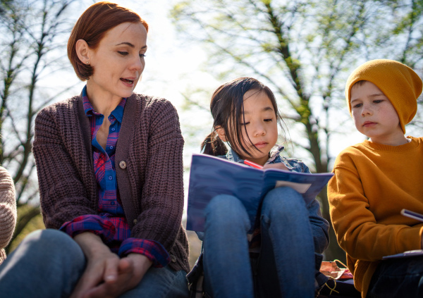 A teacher with small children sitting outdoors in city park, learning group education concept.