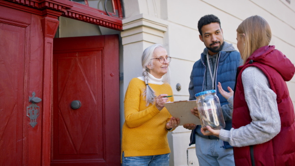 Young door to door fundraisers talking to senior woman and collecting money for charity in a street.