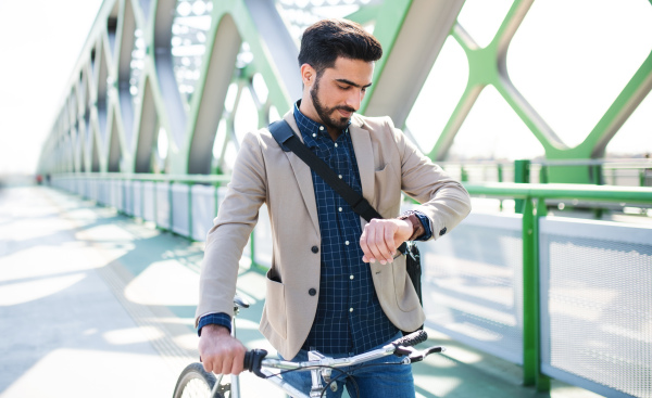 Portrait of young business man commuter with bicycle going to work outdoors in city, checking the time.