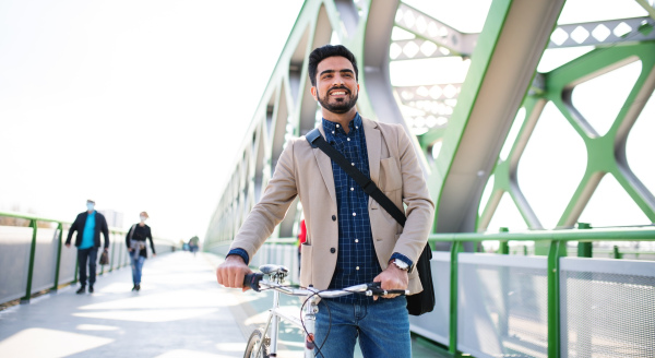 A young business man commuter with bicycle going to work outdoors in city, walking on bridge.