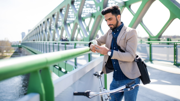 Portrait of young business man commuter with bicycle going to work outdoors in city, using smartphone.