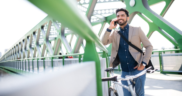 Portrait of young business man commuter with bicycle going to work outdoors in city, using smartphone.