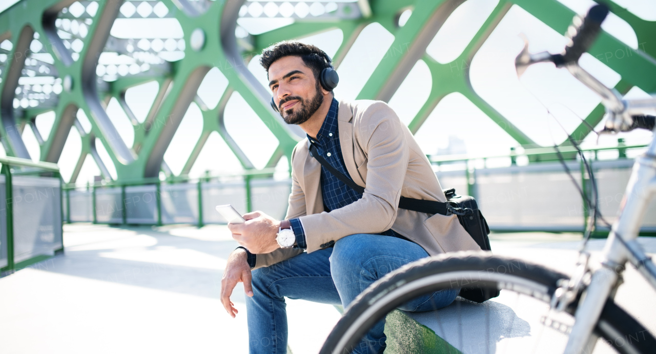 Portrait of young business man commuter with bicycle on the way to work outdoors in city, using smartphone.