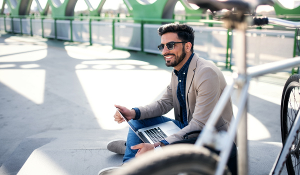 Portrait of young business man commuter with bicycle on the way to work outdoors in city, using laptop.