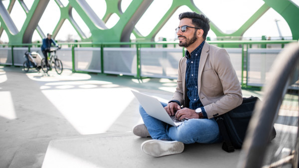 Portrait of young business man commuter with bicycle on the way to work outdoors in city, using laptop.