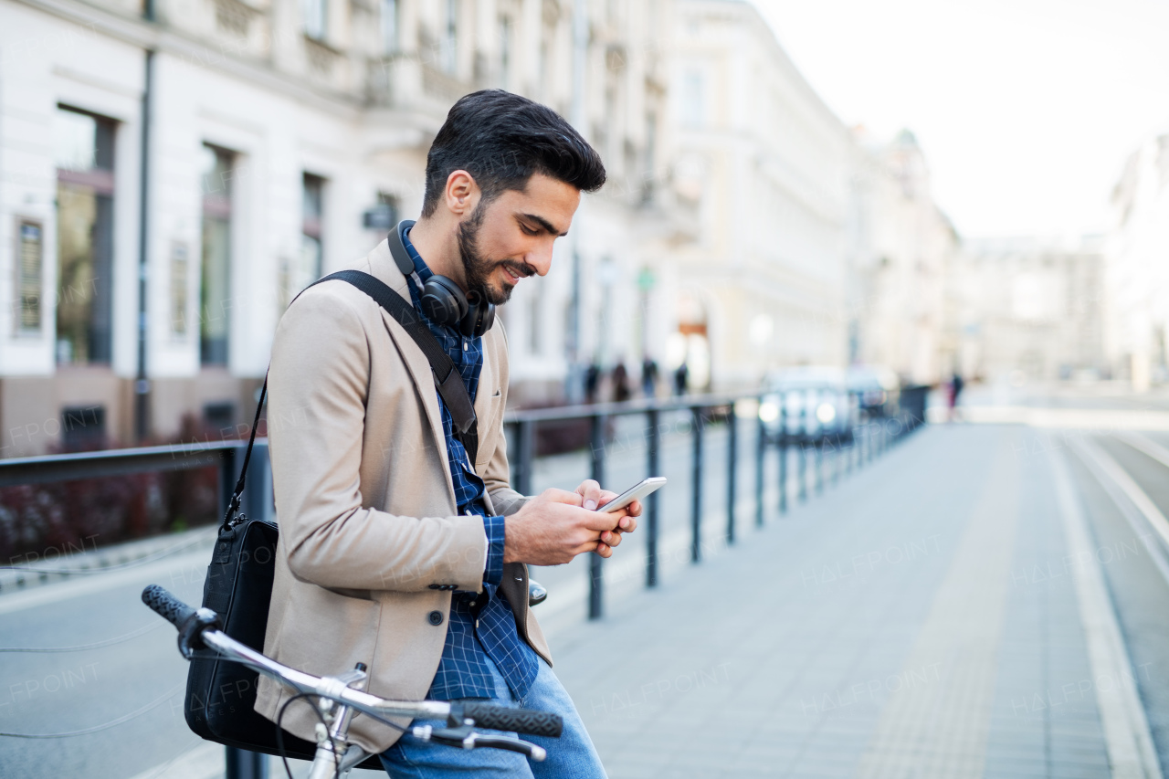 Portrait of young business man commuter with bicycle going to work outdoors in city, using smartphone.