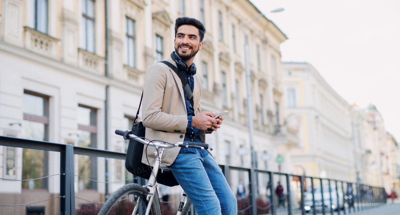 Portrait of young business man commuter with bicycle going to work outdoors in city, using smartphone.