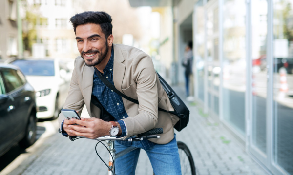 Portrait of young business man commuter with bicycle going to work outdoors in city, using smartphone.