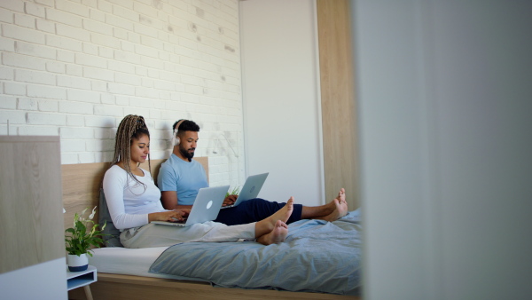 A happy multiracial young couple in pajamas sitting in bed and using laptops indoors at home.