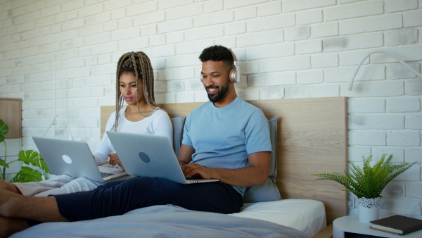 A happy multiracial young couple in pajamas sitting in bed and using laptops indoors at home.