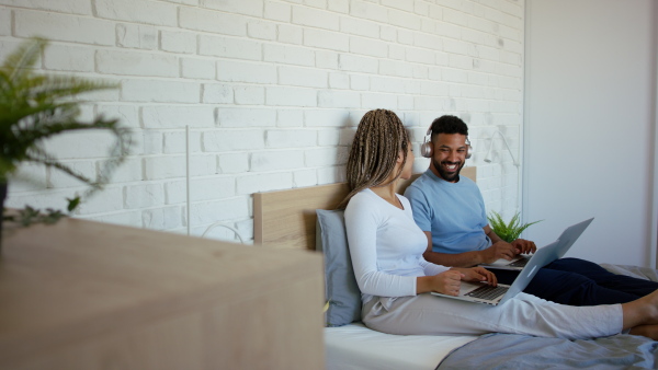 A happy multiracial young couple in pajamas sitting in bed and using laptops indoors at home, social network concept.