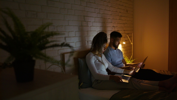 A happy multiracial young couple in pajamas sitting in bed and using laptops indoors at home at night.