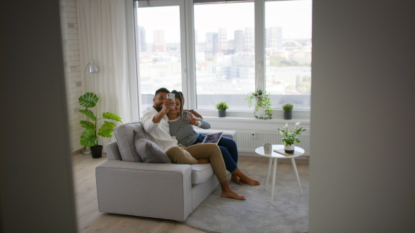 A happy multiracial young couple sitting on sofa and taking selfie indoors at home, social networks concept.