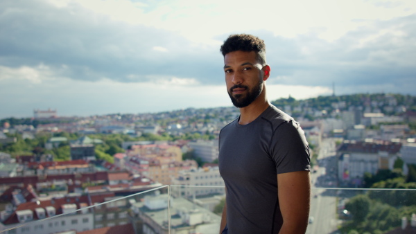 A young sport man standing with arms crossed and looking at camera on balcony outdoors in city.