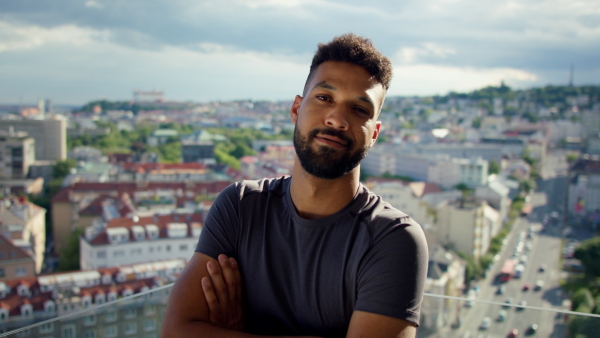 A young sport man standing with arms crossed and looking at camera on balcony outdoors in city.