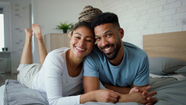 A multiracial young couple in love lying on bed, cuddling together and looking at camera indoors at home.