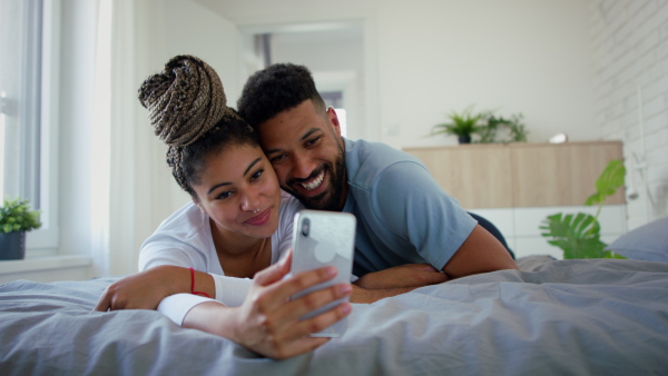 A multiracial young couple in love lying on bed and taking selfie indoors at home.