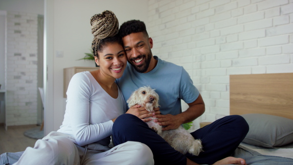 A multiracial young couple in love sitting on bed and playing with dog together indoors at home, looking at camera.