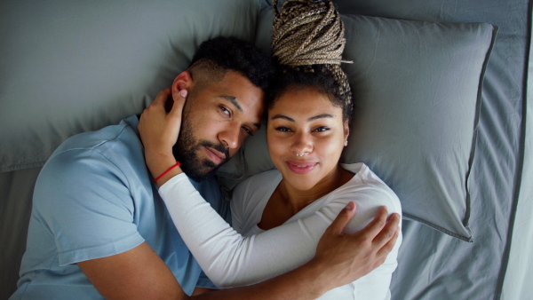 A top view of multiracial young couple in love lying on bed and cuddling together indoors at home, looking at camera.