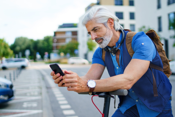 Portrait of mature man commuter with electric scooter outdoors in city, using smartphone.