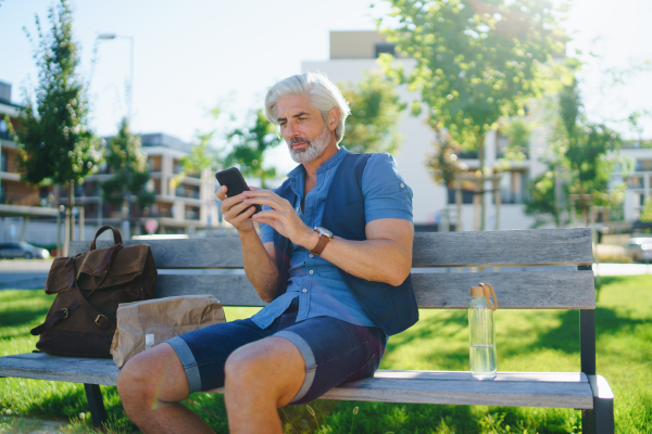 Portrait of handsome mature man sitting outdoors on bench in city, using smartphone.