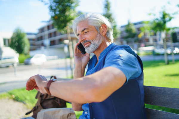 Portrait of handsome mature man sitting outdoors in city, using smartphone.