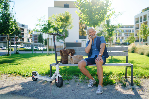 Portrait of mature man with electric scooter sitting on bench outdoors in city, using smartphone.