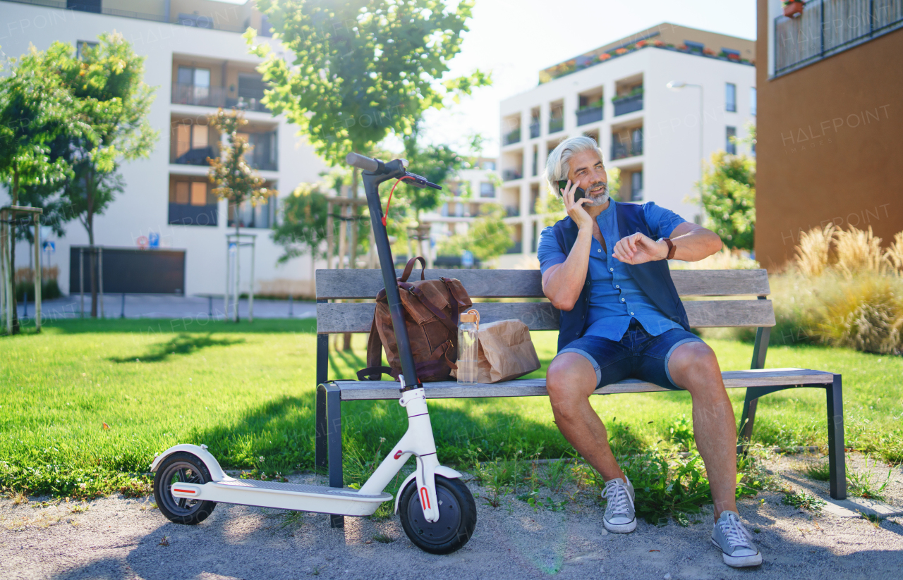 Portrait of mature man with electric scooter sitting on bench outdoors in city, using smartphone.