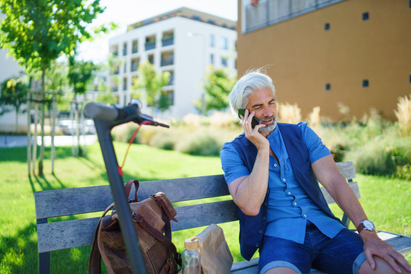 Portrait of mature man with electric scooter sitting on bench outdoors in city, using smartphone.