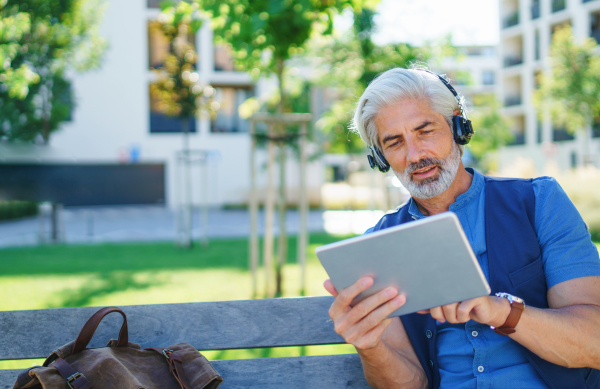 Portrait of mature man with headphones sitting outdoors in city, using tablet. Copy space.