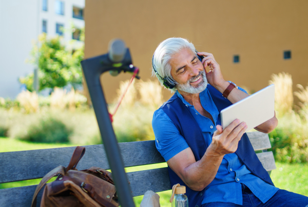 Portrait of mature man with headphones sitting outdoors in city, using tablet. Copy space.