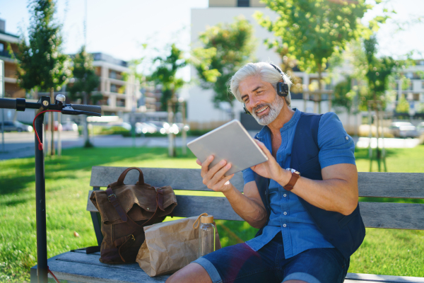 Portrait of mature man with headphones sitting outdoors in city, using tablet. Copy space.